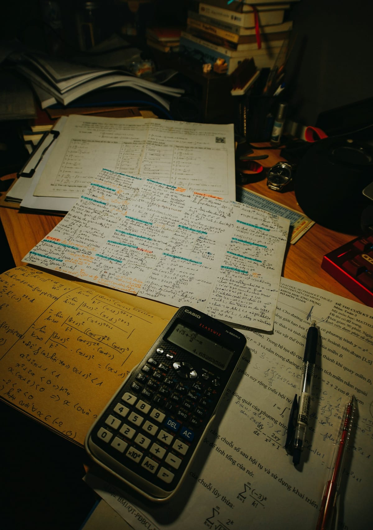 A desk with a calculator, pens and papers under a lamp.