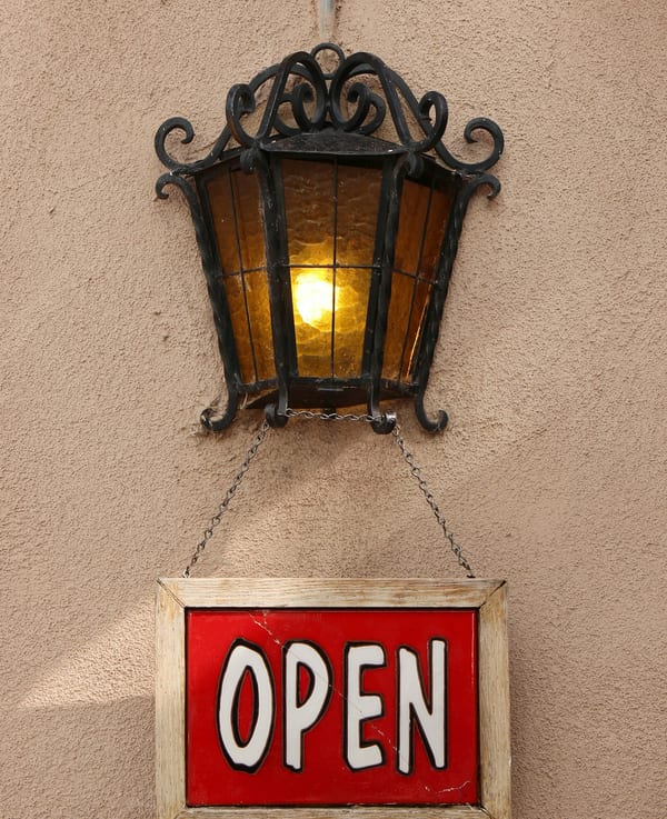 An open sign hanging under a lamp beside a shop door.
